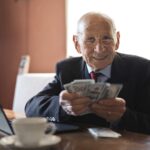Confident senior businessman holding money in hands while sitting at table near laptop