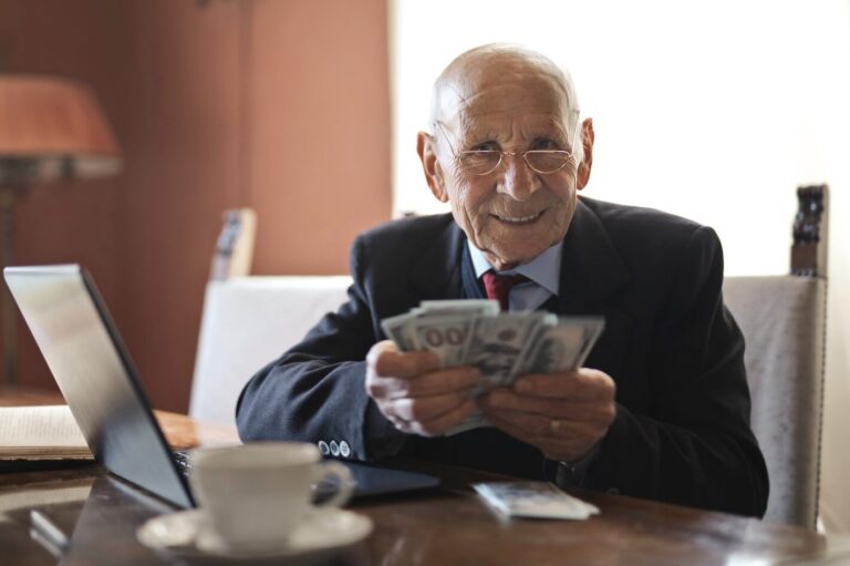 Confident senior businessman holding money in hands while sitting at table near laptop